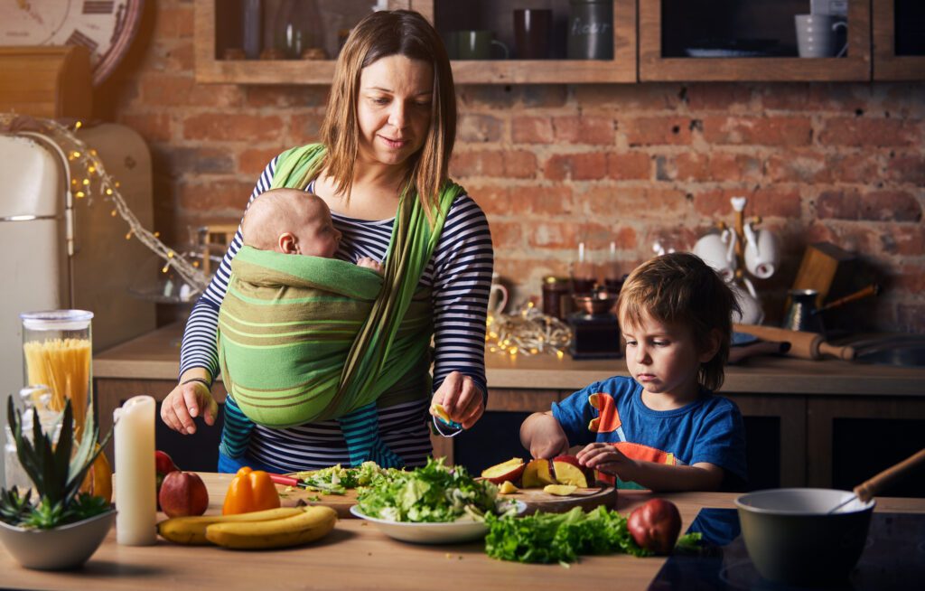 a mother with a baby in a sling, in a rustic kitchen, cooking with a preschooler